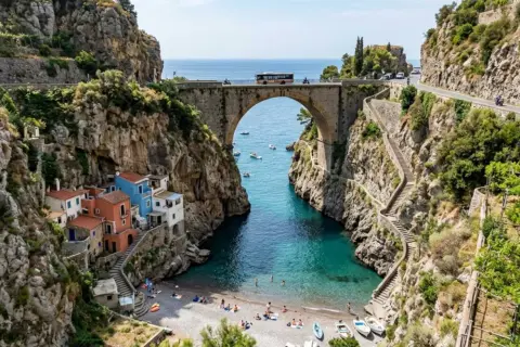 Un ponte ad arco domina un fiordo con case colorate aggrappate alla roccia. Il mare turchese invita a un bagno, mentre la strada panoramica, teatro del furore costiera amalfitana, accoglie bus e moto.