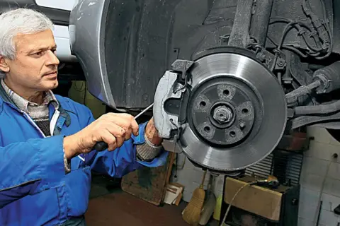Mechanik w niebieskim kombinezonie naprawia hamulce, wymieniając klocki. W tle widać elementy układu kierowniczego i pompowtryski.