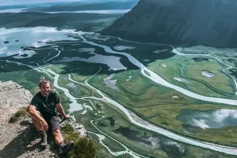 Man vilar på en klippa med utsikt över Sarek nationalparks meandrande vattendrag och fjällandskap.