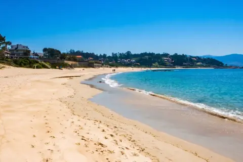 La **playa pintens** se extiende bajo un cielo azul intenso, con aguas turquesas acariciando la arena dorada. Casas y vegetación salpican la costa.