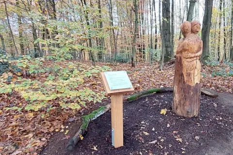 Holzschnitzerei zweier sich umarmender Figuren neben einem Schild mit der Aufschrift "Fatima Kapelle" im Herbstwald.