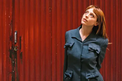 Catalina Sopelana, con su cabello rojizo y mirada perdida, posa frente a una puerta roja acanalada, luciendo un traje gris con detalles de lazos.
