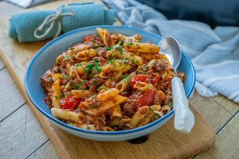 Herzhafte Nudeln mit Hackfleisch und Tomaten, garniert mit frischer Petersilie, serviert in einer blauen Schüssel auf einem Holzbrett.