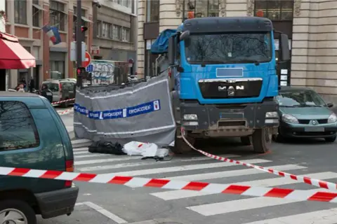 Scène d'accident avec camion bleu, voiture verte et ruban de sécurité. Un autocollant angle mort Europe est visible sur le camion.