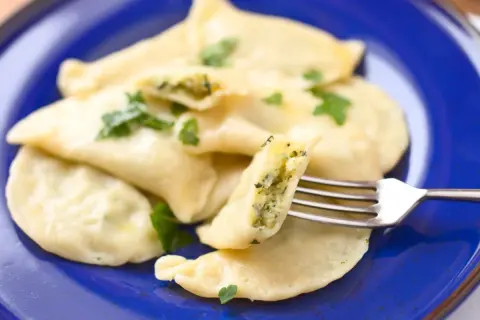 Pyszne pierogi ze szpinakiem i serem, udekorowane natką pietruszki, na niebieskim talerzu. Jedna pierożka jest nadziana widoczna na widelcu.