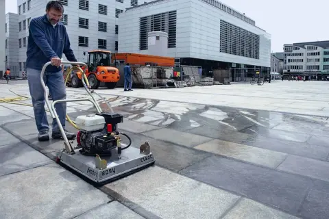 Uomo pulisce autobloccanti con idropulitrice in una piazza. L'acqua riflette la luce sui blocchi di pietra.