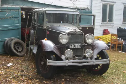 En gammal Dodge-bil står parkerad utanför ett garage. Bilen är mörkt vinröd med kromdetaljer och har en klassisk design.