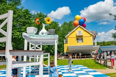 Dzieci bawią się w surrealistycznym parku, gdzie dom unosi się na balonach, a stół jest gigantyczny. Idealne miejsce na rodzinne przygody w parkach dla dzieci.