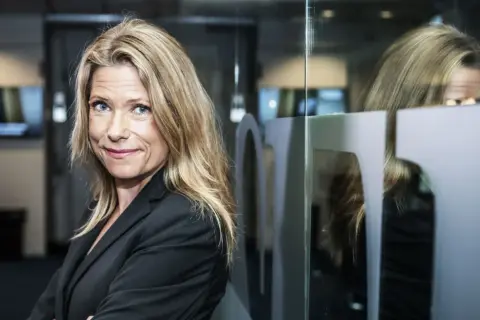 Petra Nordlund, med sitt blonda hår och blå ögon, ler mot kameran. Hon bär en svart kavaj och står framför en glasvägg med en spegelbild.