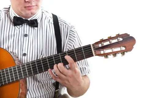 Mężczyzna w muszce i koszuli w paski gra na gitarze, trzymając palce na gryfie. Widoczne są tabulatury na gitarę.
