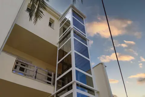 Moderno montacarichi persone esterno in vetro e metallo bianco, che sale lungo un edificio color crema, con cielo nuvoloso al tramonto.