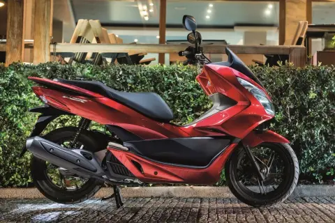 Czerwony skuter Honda PCX, jeden z popularnych **marek skuterów**, zaparkowany przed zielonym żywopłotem.