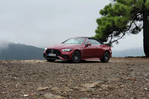 Czerwony kabriolet Mercedes-AMG CLE 53 na kamienistym podłożu, z drzewem i mglistymi górami w tle.