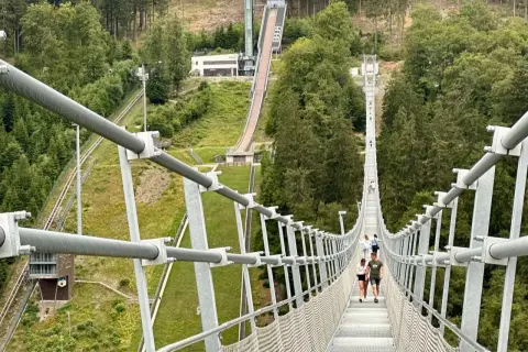 Menschen spazieren über den Willingen Skywalk, eine Hängebrücke mit Blick auf eine Skisprungschanze und bewaldete Hänge.