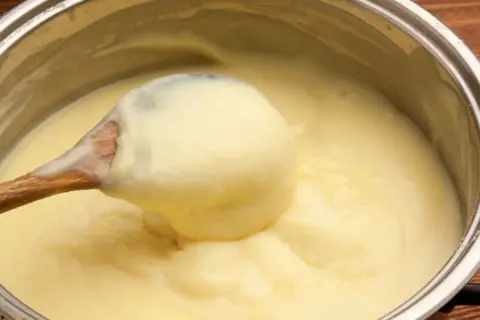 Une cuillère en bois prélève une généreuse portion de crème pâtissière onctueuse dans une casserole.