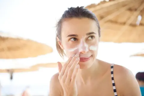 Frau trägt Sonnencreme auf, um Pickel durch Sonne zu vermeiden. Sie ist am Strand unter Sonnenschirmen.