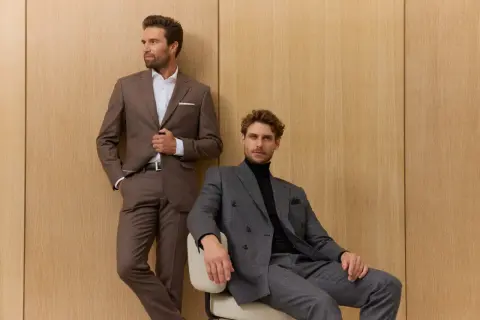 Zwei Männer in eleganten Anzügen vor Holzvertäfelung. Der eine steht, der andere sitzt. Die perfekte sakko passform unterstreicht ihre maskuline Silhouette.