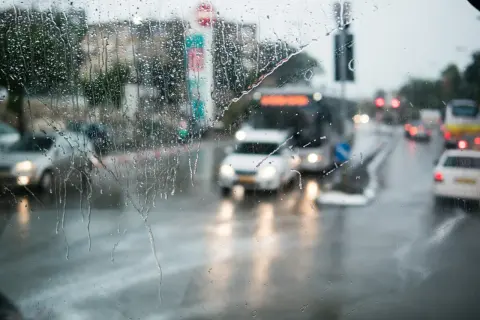 Gouttes de pluie sur un pare-brise, floutant la circulation urbaine. Les feux position seuls des voitures et des bus créent des traînées lumineuses sur la chaussée mouillée.
