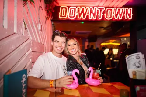 Coppia sorridente in un locale di Londra, con cocktail rosa e un'insegna al neon "DOWNTOWN".