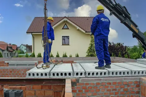 Dwóch budowlańców w niebieskich kombinezonach montuje pustaki stropowe. Dźwig podnosi kolejne elementy.