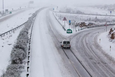Nieve en la sierra de Madrid hoy. Una furgoneta blanca circula por una carretera nevada, con señales de tráfico y vegetación cubierta de blanco.