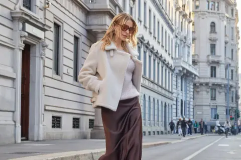 Donna con tailleur in inverno: un cappotto corto in montone bianco sopra un maglione e gonna di seta marrone.