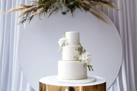 Eleganckie torty weselne wzory: biały, trzypiętrowy tort z delikatnymi kwiatami i koronkowym zdobieniem, prezentowany na złotym postumencie.