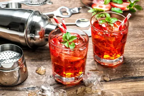 Zwei rote Cocktails mit Erdbeeren und Minze, Eiswürfel, Cocktailshaker und Barzubehör auf Holztisch.