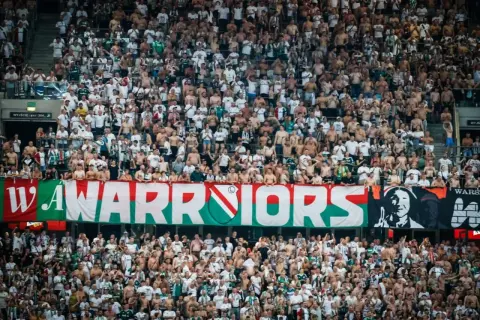 Tłum kibiców Legii Warszawa, z transparentem "WARRIORS" i herbem klubu. Wszyscy w biało-zielonych barwach, gotowi do dopingu.