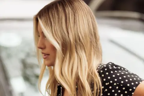 Perfil de mujer con cabello rubio mantequilla, ondas suaves y un vestido negro con lunares blancos.