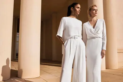 Due donne eleganti in bianco, un'ispirazione su come vestirsi ad un matrimonio con pantaloni ampi o abito longuette.