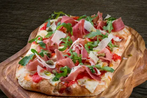 En avlång pinsa pizza med prosciutto, ruccola och parmesan på en träbricka.
