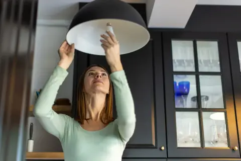 Mujer cambia bombilla en lámpara. ¿Se puede vender una casa sin luz ni agua? Quizás, pero con luz es más fácil.