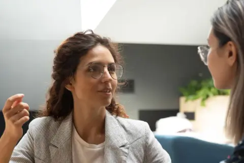 Två kvinnor i samtal, en med glasögon och lockigt hår, den andra med långt hår. En man syns i bakgrunden. Scenen utstrålar autentiskt ledarskap genom öppen kommunikation.