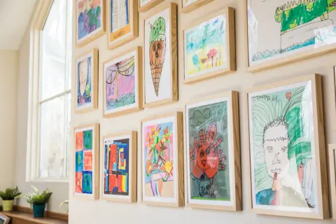A vibrant kids art gallery wall showcases colorful drawings of ice cream, flowers, and portraits, each framed in light wood.
