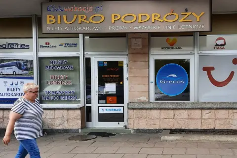 Biuro podróży z ofertą Grecos. Kobieta w paski idzie chodnikiem przed witryną z logo Grecos.
