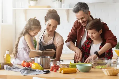 Famille préparant un repas. Le père aide son fils à couper des légumes. La mère et la fille consultent un livre de recettes. On se demande : lactose c'est quoi ?