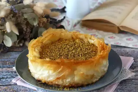 En gyllene baklava cheesecake med pistagenötter, serverad på en grå tallrik. En vacker dessert att njuta av med en god bok.