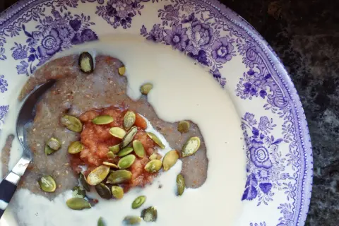 En skål med skrädmjölsgröt, toppad med äppelmos och pumpafrön, serverad i en vacker lila-vit tallrik.