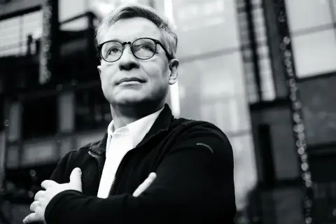 Mężczyzna w okularach, z rękami skrzyżowanymi na piersi, patrzy w górę. Jego postawa sugeruje, że jest alior ostrożny, analizując sytuację.