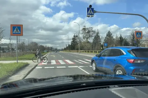 Niebezpieczne wyprzedzanie na pasach na drodze dwupasmowej. Niebieski SUV omija rowerzystkę stojącą przy przejściu.