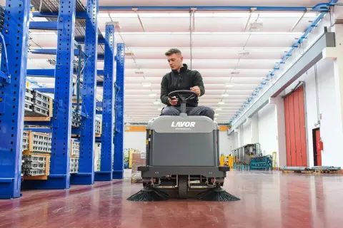 Hombre operando una maquina limpieza industrial Lavor en un almacén con estanterías azules.