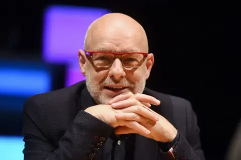 Brian Eno, eno musicista inglese, con occhiali rossi e mani intrecciate.