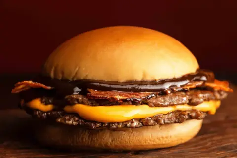 Gros plan sur un burger double viande, fromage fondant, bacon croustillant et sauce barbecue, sur une planche en bois.