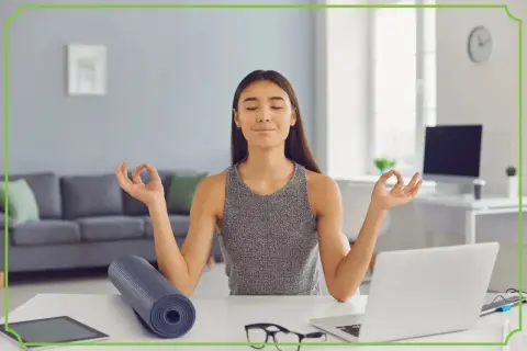 Kobieta medytuje przy biurku, osiągając work life balance. Obok leży mata do jogi i laptop.