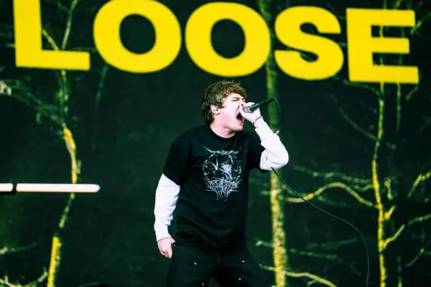 Wokalista z otwartymi ustami śpiewa do mikrofonu na tle sceny z napisem "LOOSE".