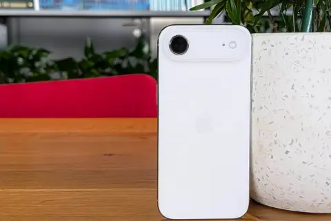 Biały iPhone 5c z charakterystycznym modułem aparatu, stojący na drewnianym blacie obok doniczki.