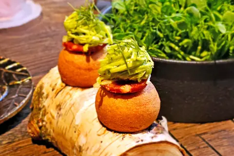 Dwa apetyczne mini-burgerki z zielonymi dodatkami, serwowane na pniu brzozy. Idealne na przekąskę, jak w najlepszych **bottiglieria 1881 opinie**.