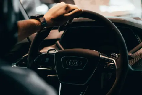 Dłoń na kierownicy Audi, z zegarkiem na nadgarstku. W tle widać fragment deski rozdzielczej, sugerujący wnętrze nowoczesnego samochodu, jak w tabeli z cenami rynkowymi samochodów używanych.