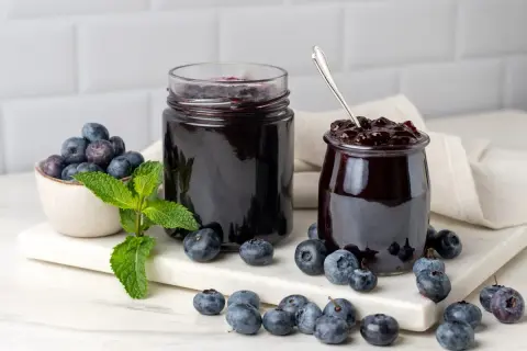 Due barattoli di marmellata di mirtilli, uno aperto con un cucchiaino, circondati da mirtilli freschi e foglie di menta.
